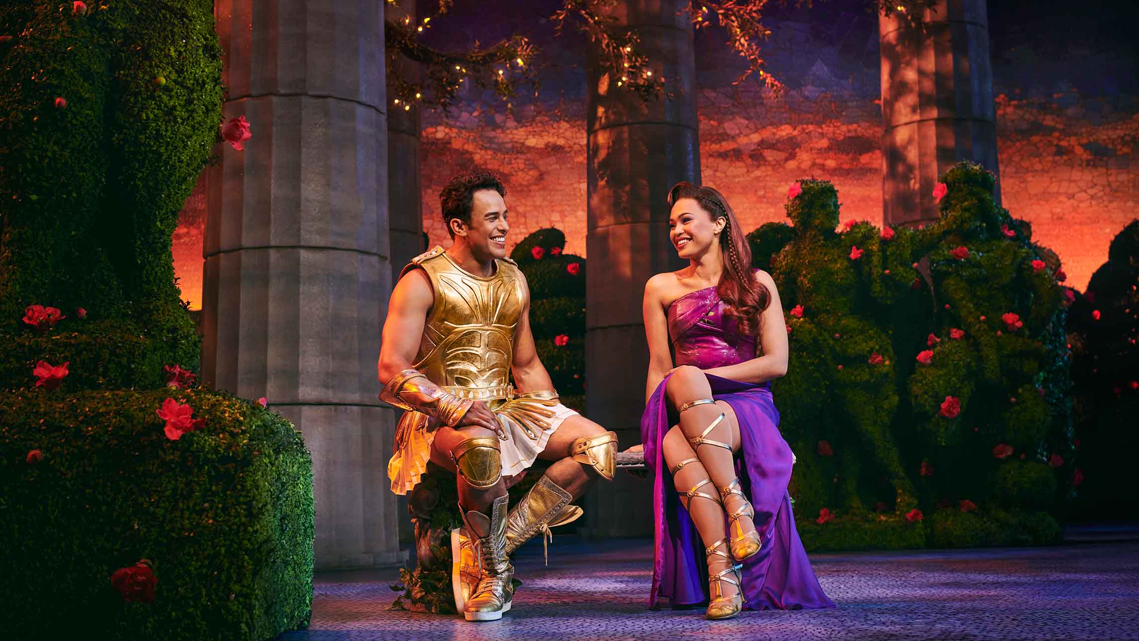 Actors Luke Brady (Hercules) and Mae Ann Jorolan (Meg) on stage in Disney's Hercules smiling at each other whilst sat down